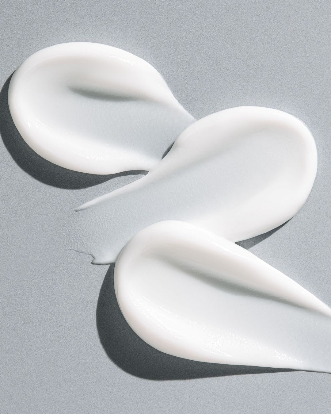 Tre strisce di crema bianca e lucida sono spalmate su uno sfondo grigio chiaro, mostrando la consistenza ricca e liscia del prodotto.