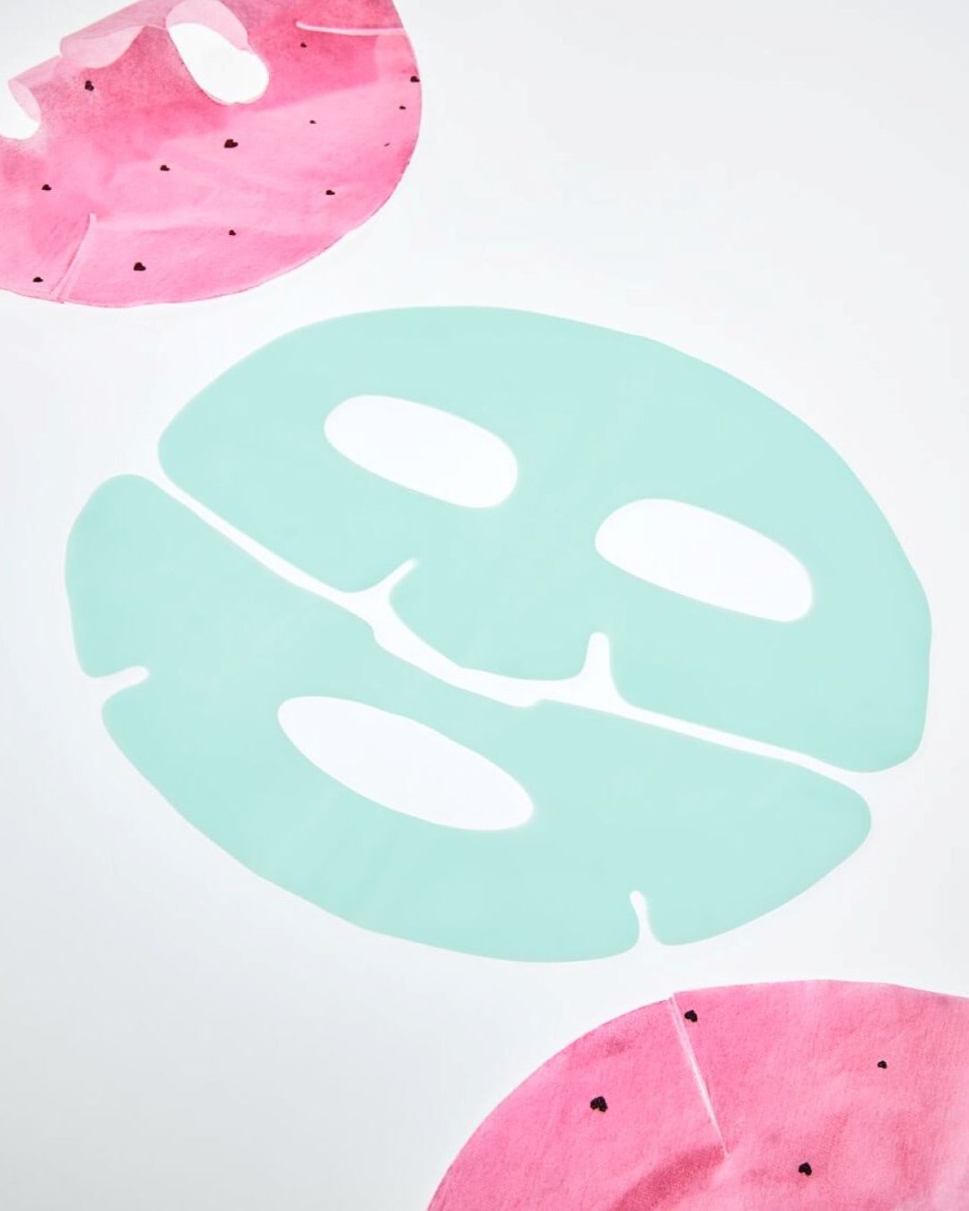 Una maschera viso verde acqua a forma di foglia di Monstera è posizionata al centro, affiancata da due maschere viso rosa a forma di anguria con piccoli semi neri, su uno sfondo bianco.