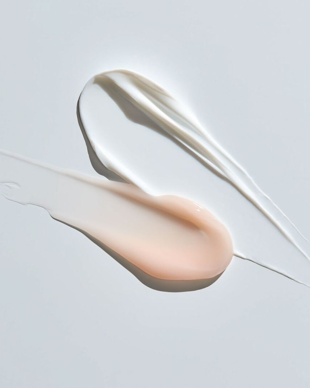 Due strisce di crema, una bianca e una color pesca, sono spalmate su una superficie bianca, mostrando la loro consistenza liscia e ricca.