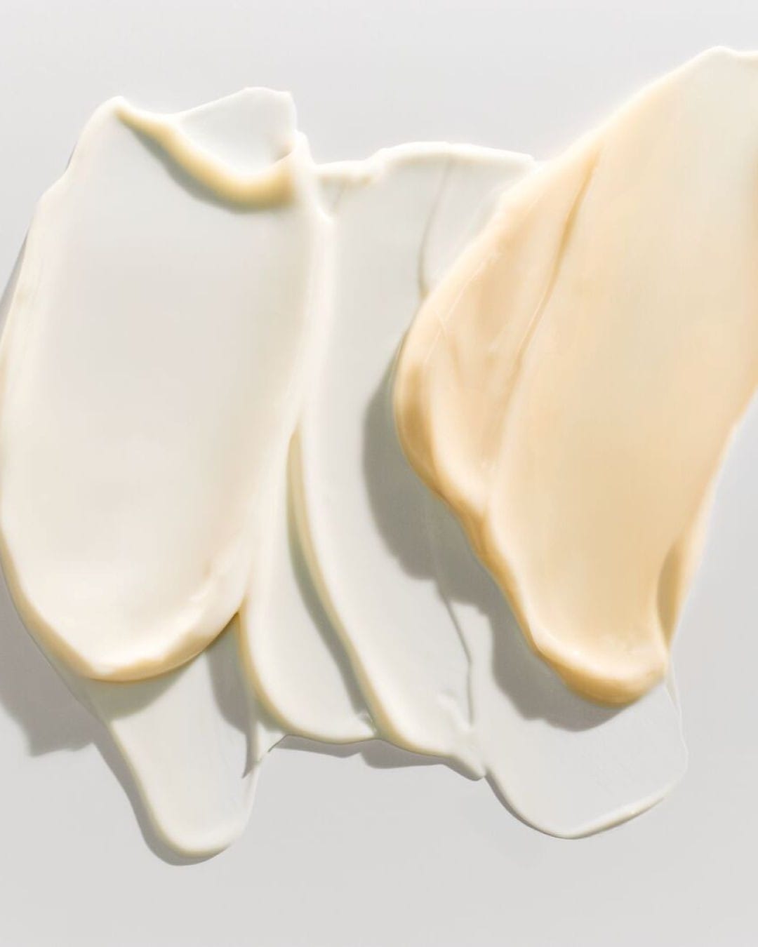 Una serie di creme e lozioni per la cura della pelle, con texture ricche e colori che vanno dal bianco al beige chiaro, sono disposte su una superficie bianca, creando un effetto visivo elegante e invitante.