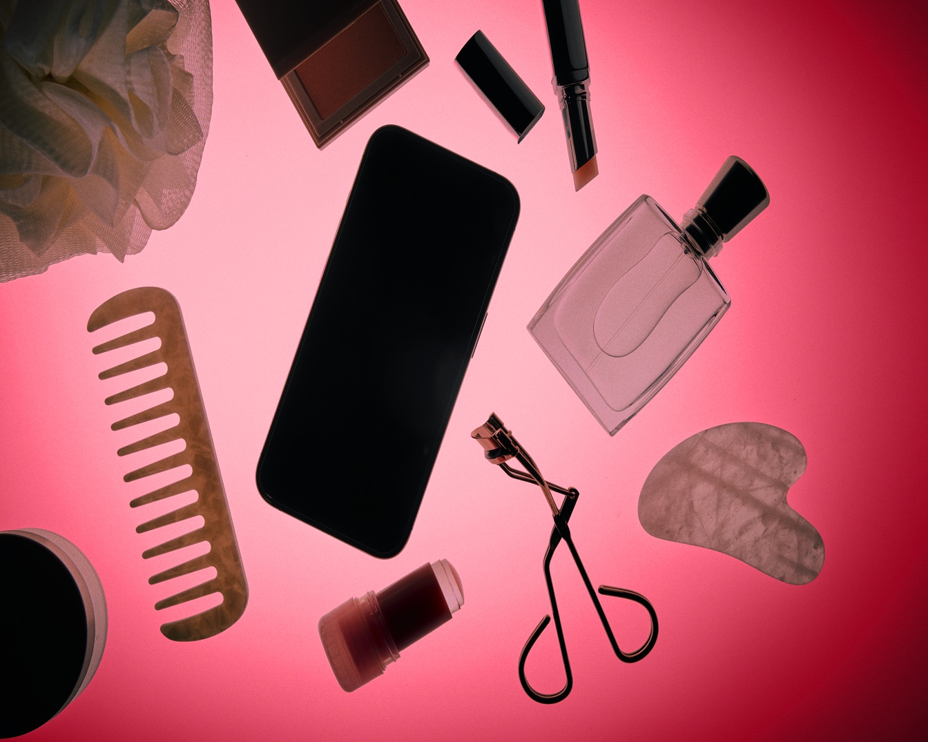 Une vue aérienne artistique présente une sélection d'accessoires de beauté et de bien-être, incluant un peigne en bois, un flacon de parfum, un recourbe-cils et un smartphone disposés sur un fond rose éclatant. Achetez vos favoris beauté dès maintenant !
