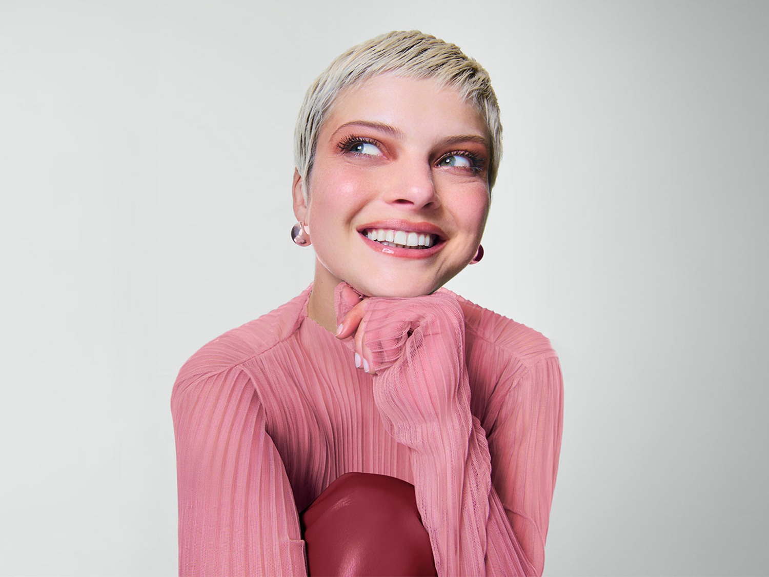 Een stralende vrouw met kort blond haar en roze oogschaduw draagt een roze plooitop en glimlacht vrolijk.