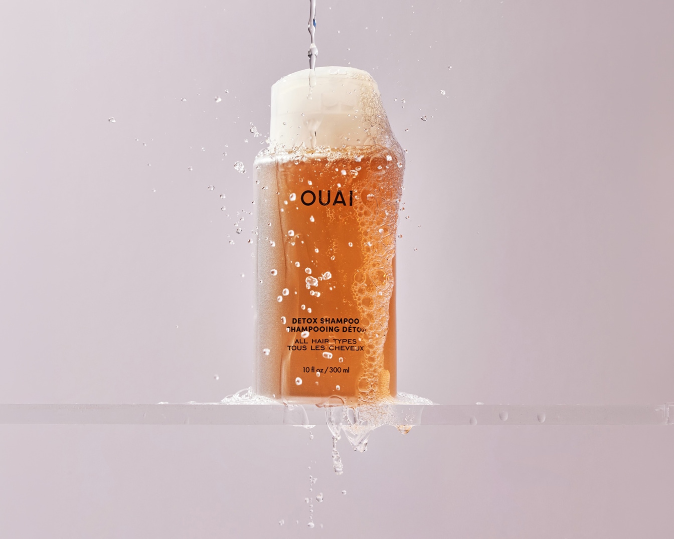 Flacon de shampoing détox OUAI de 300 ml enrobé de mousse et arrosé d'eau sur une surface claire, idéal pour tous les types de cheveux. Achetez dès maintenant pour purifier votre chevelure !
