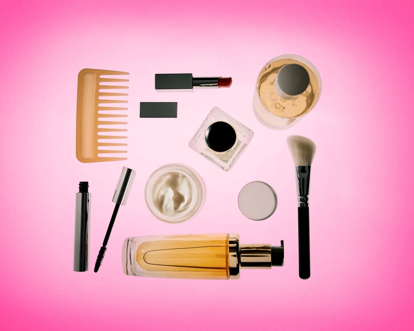 Růžové pozadí s různými kosmetickými produkty, včetně hřebenu, řasenky, rtěnky, krému, make-upu, parfému a štětce na make-up, vše úhledně uspořádané.