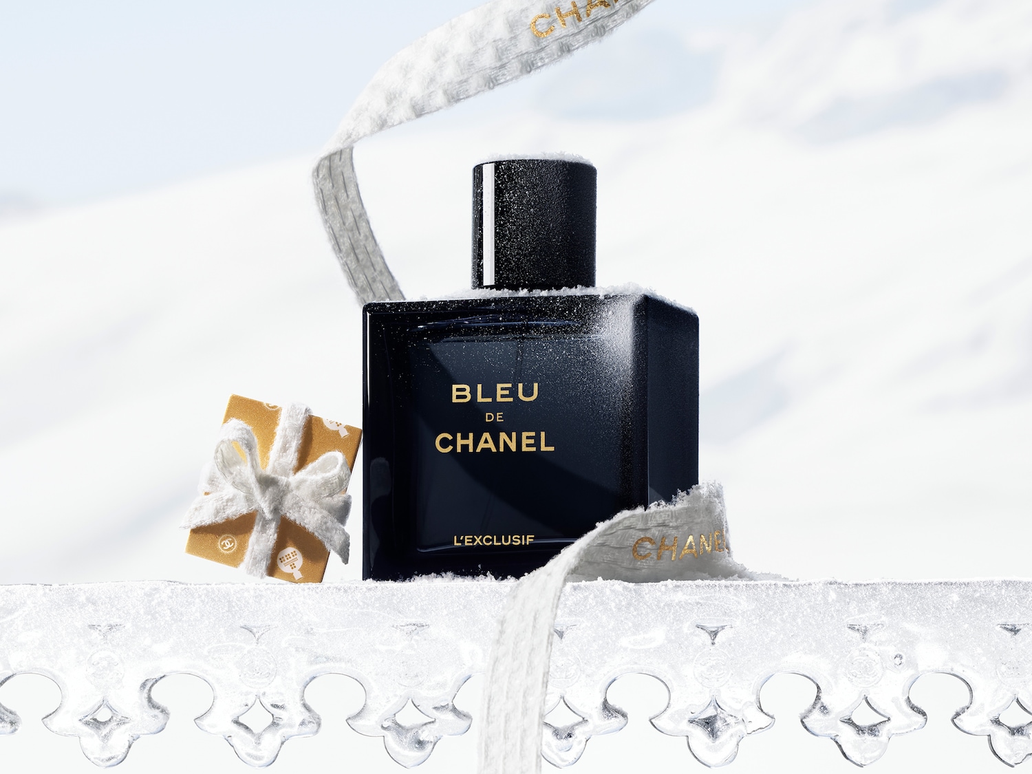 Een donkerblauwe fles Bleu de Chanel L'Exclusif staat op een besneeuwd oppervlak, omringd door een wit lint en een klein goudkleurig geschenkje.