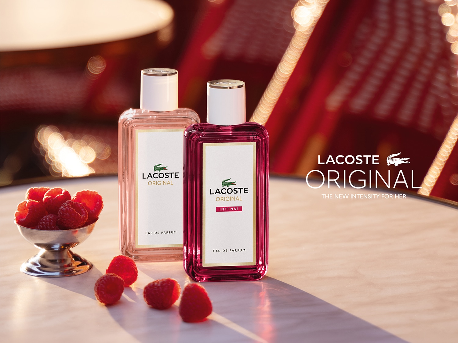Dwa prostokątne flakony perfum Lacoste Original z białymi nakrętkami stoją na stole w otoczeniu świeżych malin, promując nową intensywność zapachu dla kobiet.