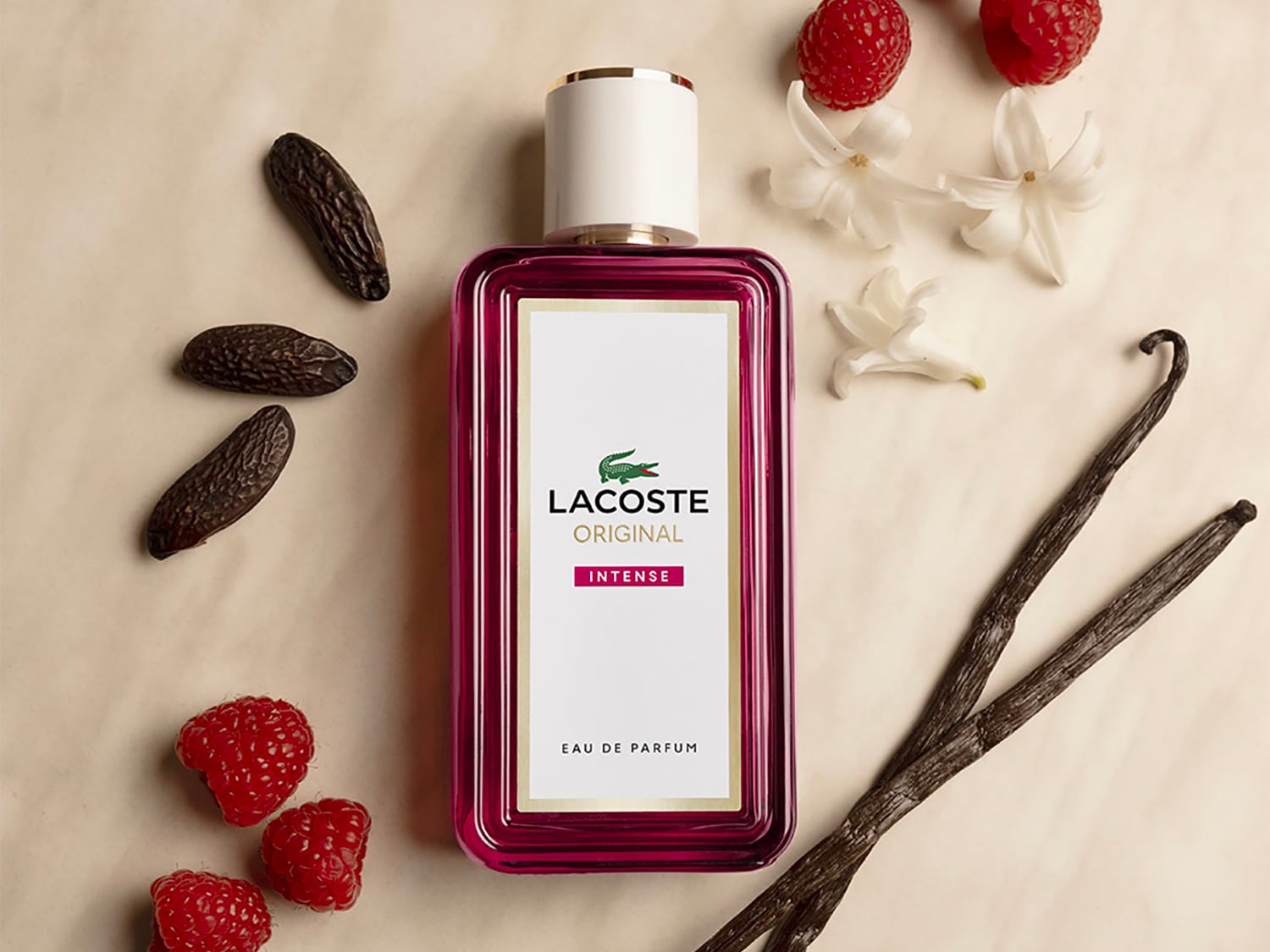 Prostokątna szklana flaszka perfum Lacoste Original Intense Eau de Parfum w głębokim różowym kolorze z białym korkiem leży na jasnej powierzchni, otoczona świeżymi malinami, wanilią, białymi kwiatami i ziarnami kakaowca. Odkryj ten wyjątkowy zapach już teraz!