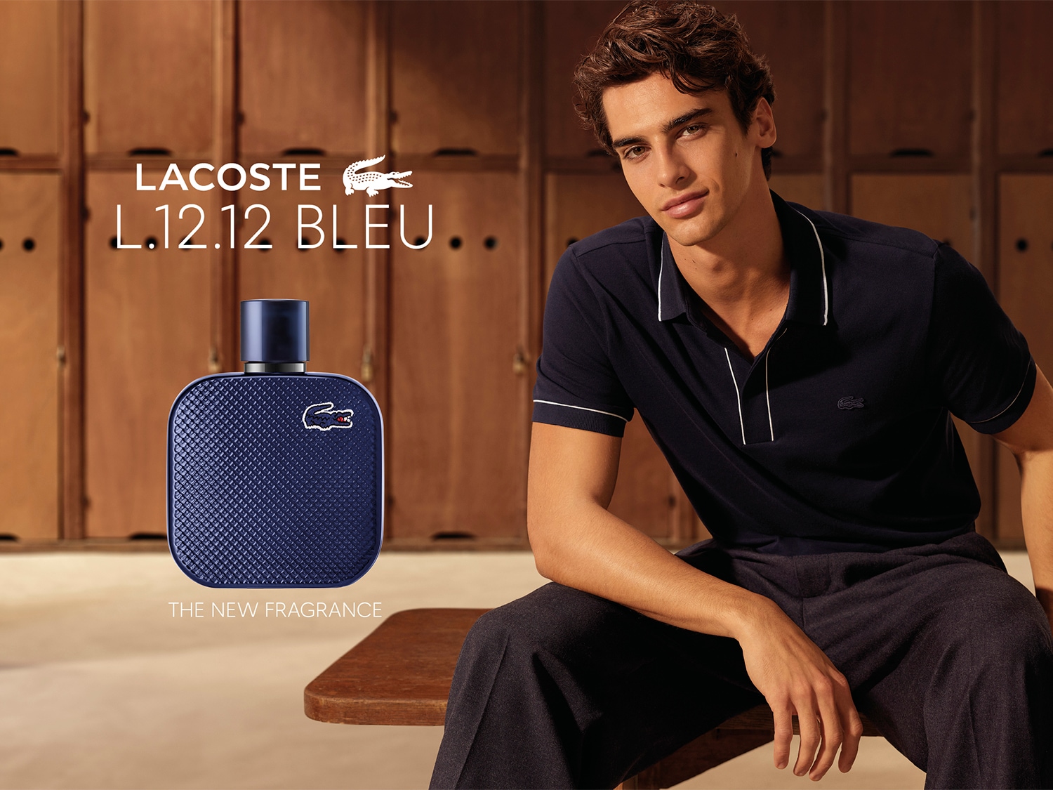 Młody mężczyzna w ciemnogranatowej koszulce polo siedzi w szatni obok niebieskiego flakonu perfum Lacoste L.12.12 Bleu, odkryj nowy zapach już teraz.