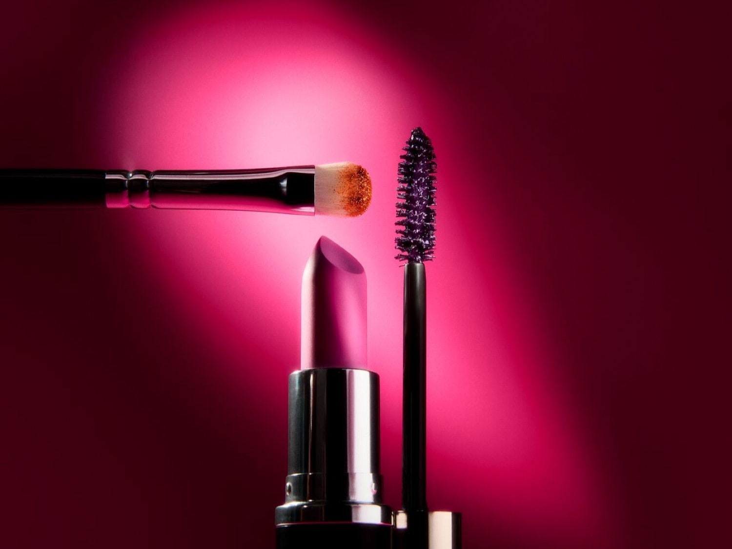 Un set di prodotti di bellezza tra cui un rossetto rosa in un elegante astuccio argentato, un pennello da trucco con setole morbide e un mascara nero con scovolino definito, il tutto su uno sfondo rosa vibrante.