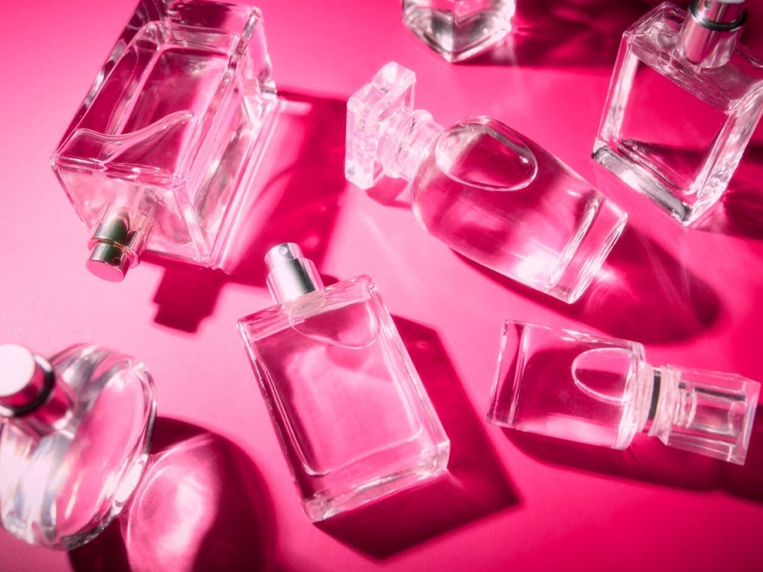 Diverse bottiglie di profumo in vetro trasparente di varie forme sono disposte ordinatamente su una superficie di colore rosa vibrante.