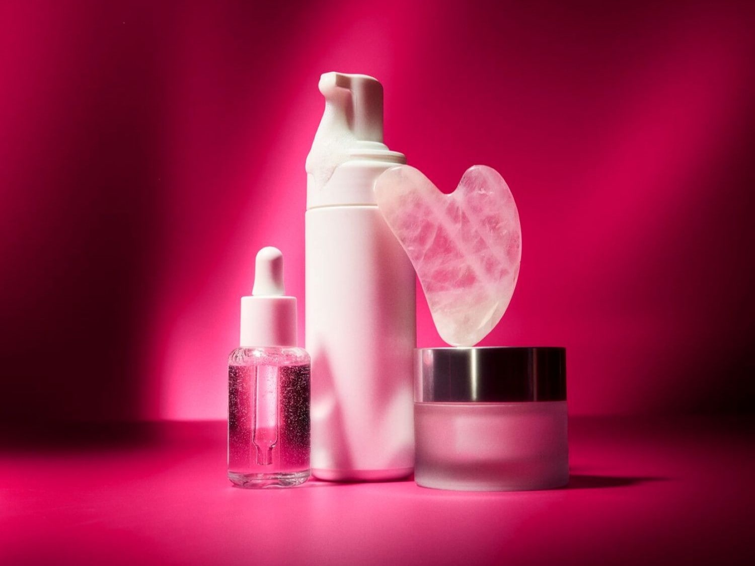 Una selezione di prodotti di bellezza su sfondo rosa, tra cui un flacone trasparente con contagocce, un erogatore bianco e un vaso di crema con pietra Gua Sha. Acquista ora per completare la tua routine di cura della pelle.