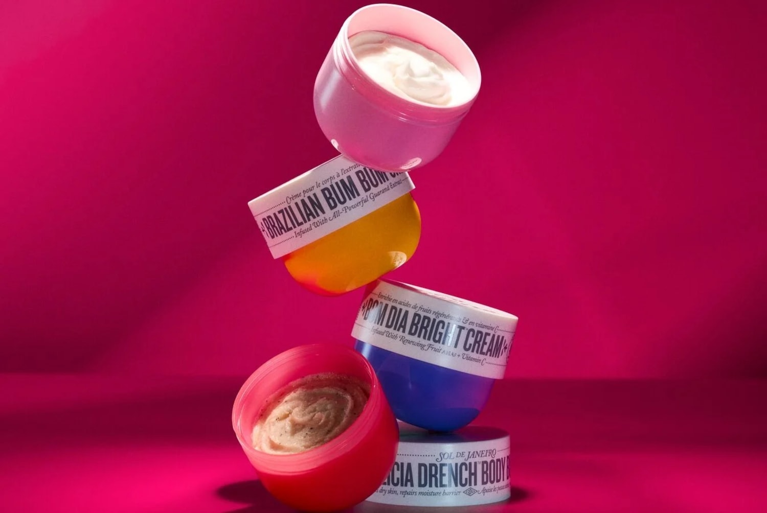 Una pila verticale di barattoli colorati Sol de Janeiro su sfondo fucsia, che mostra creme idratanti ricche e uno scrub corpo esfoliante. Acquista ora per una pelle luminosa.