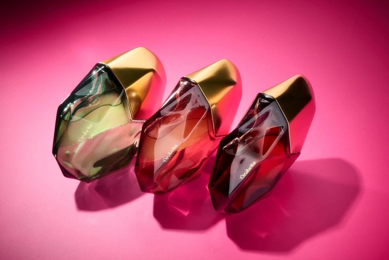 Tre flaconi di profumo Orabella con design sfaccettato e tappo dorato sono disposti su uno sfondo rosa vibrante, scopri la collezione e acquista ora.