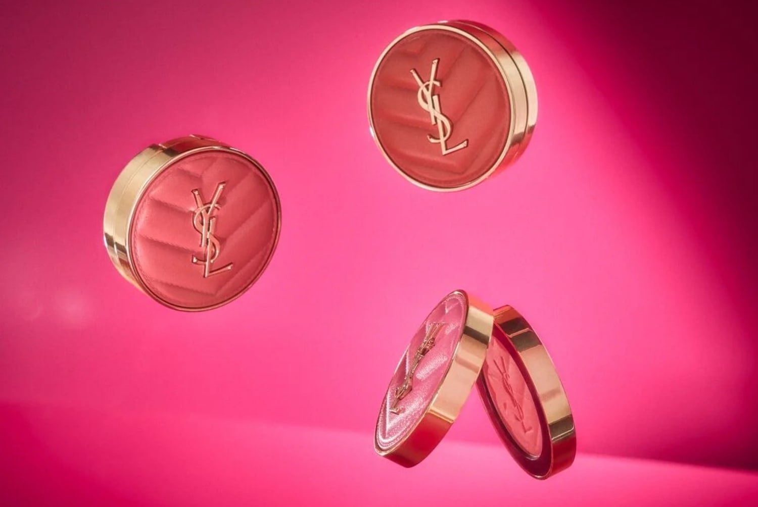 Tre eleganti confezioni rotonde di Yves Saint Laurent in tonalità rosa con dettagli dorati e logo in rilievo fluttuano su uno sfondo rosa vivace.