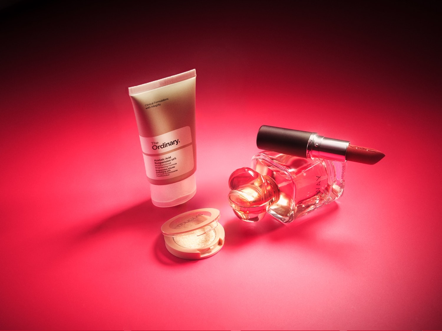 Een stijlvolle opstelling van beautyproducten op een rode achtergrond, waaronder een tube The Ordinary huidverzorging, een open Douglas highlighterdoosje, een flesje parfum en een rode lippenstift.