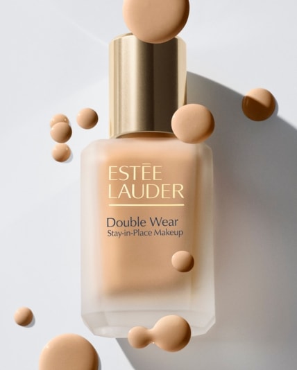 Ein Fläschchen „Estée Lauder Double Wear Stay-in-Place Makeup“ in mattem Glas mit goldenem Verschluss steht vor hellem Hintergrund, umgeben von schwebenden Tropfen in verschiedenen Hauttönen.