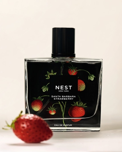 Eine klare Glas-Parfümflasche mit der Aufschrift „NEST New York Santa Barbara Strawberry Eau de Parfum“, verziert mit Illustrationen von Erdbeeren und grünen Blättern, steht auf einer hellen Oberfläche; im Vordergrund liegt eine frische Erdbeere.