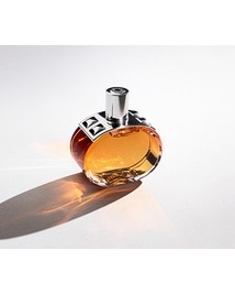 Hermès Parfümflakon aus transparentem Glas mit bernsteinfarbener Flüssigkeit. Der runde Flakon mit silberfarbenem Verschluss steht vor hellem Hintergrund. Warmes Licht erzeugt einen langen Schatten und orangefarbene Lichtreflexe auf der Oberfläche.
