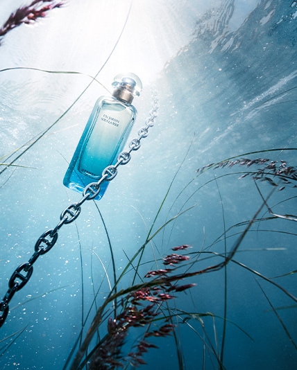 Türkisfarbener Hermès-Parfümflakon ‚Un Jardin Sous la Mer‘ schwebt unter Wasser an einer Kette, umgeben von Seegras und hellem Lichteinfall.